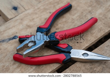 two pairs of red and black handled pliers, including a standard combination plier and a needle-nose plier, rest crosswise on top of rough wooden pallet boards.
