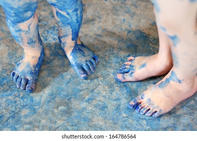 two pairs of very messy blue painted kid's feet are standing on a floor covered in spilled paint