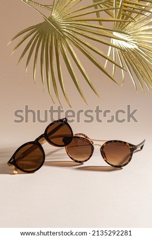 Two pairs of women sunglasses on a beige background with golden palm leaves. Minimalism, summer fashion concept. Trendy sunglasses still life. Optic store discount, sale. Close up, vertical