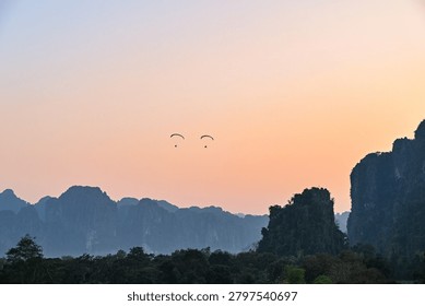 Two paragliders soar above the dramatic karst mountain landscape of Luang Prabang, Laos, during sunset. The image captures the beauty of Southeast Asia’s natural scenery, combining adventure tourism, rugged limestone cliffs, and a peaceful evening sky