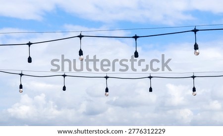 Two parallel strands of vintage-style black bistro string lights hang against a backdrop of a partly cloudy blue sky. 