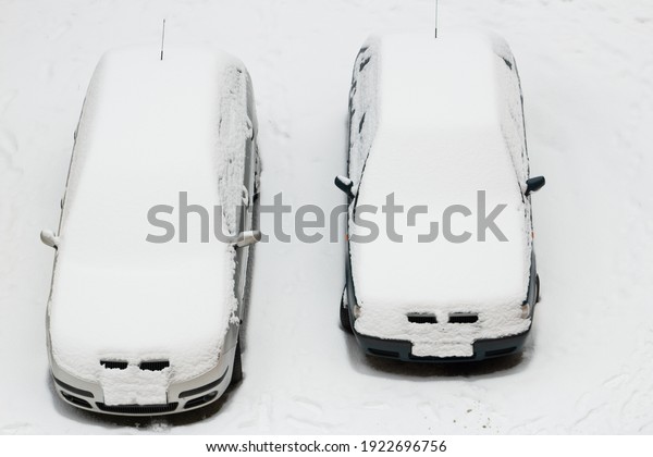 Two parked cars stand in a snowy parking lot and\
are covered with snow. A snowstorm struck. The cars and roads are\
under the snow.