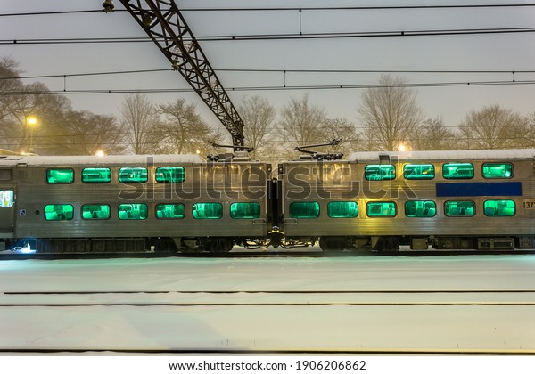 Two passenger train cars at rest\
in the snow in the evening on overcast night in urban\
Chicago