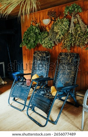 Two patterned reclining chairs with colorful towels and felt sauna hats rest in a traditional Latvian steam room. Herbal oak and plant bundles hang on the wooden wall