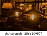 Two pendant lamps in a dimly lit room hanging from a black metal fixture. Night café interior. Warm light. Defocused interior of restaurant or bar, with tables and chairs visible.