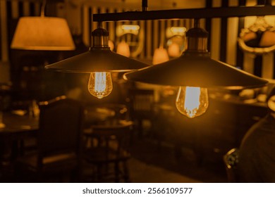 Dos lámparas colgantes en una habitación poco iluminada colgando de un accesorio de metal negro. Interior del café nocturno. Luz cálida. Interior desenfocado de restaurante o bar, con mesas y sillas visibles.