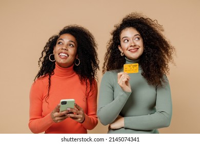 Two Pensive Young Curly Black Women Friends 20s In Casual Shirts Clothes Using Mobile Cell Phone Hold Credit Bank Card Look Up Lost In Thought Isolated On Plain Pastel Beige Background Studio Portrait