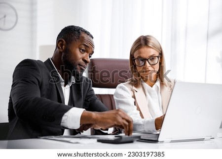 Two people analyze documents on a laptop while sitting at a table in the office. Business partners at work in the office are discussing projects. Multi-racial collaboration.