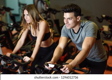 Two People Biking In The Gym, Exercising Legs Doing Cardio Workout Cycling Bikes. Couple In A Cyclo Indoor Class Wearing Sportswear.