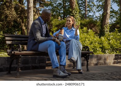 Two people in business clothes, a blonde white woman and a bald black man having a conversation on a park bench and enjoying takeaway coffees. - Powered by Shutterstock