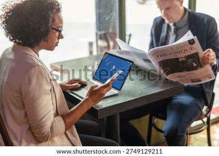 Two people at a cafe table; one uses a smartphone and laptop, the other reads a newspaper. They appear engaged in their activities.