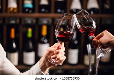 Two people clinking with glasses of red wine, celebrating success or speaking toast in wine restaurant, against racks with wine bottles, close up - Powered by Shutterstock