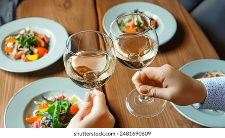 Two people clinking their glasses of Wine. The photo is taken from above, showcasing their plates set on a wooden table.  The atmosphere suggests a fine dining experience. - Powered by Shutterstock