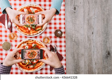 Two People Eating A Vegetable Pizza At The Restaurant, They Are Taking Pictures Of Food With Their Smartphones