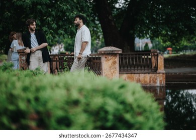 Two people engaged in conversation while standing near a bridge in a peaceful park. The lush greenery, water reflections, and relaxed stance indicate a friendly interaction in an outdoor environment.