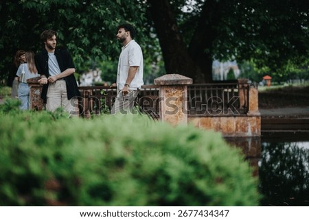 Two people engaged in conversation while standing near a bridge in a peaceful park. The lush greenery, water reflections, and relaxed stance indicate a friendly interaction in an outdoor environment.