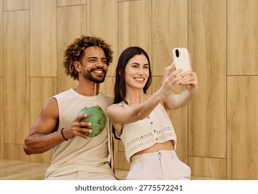 Two people enjoy coconuts while sitting in water at a pool area and using a smartphone camera  taking a selfie photo in a warm location during summer