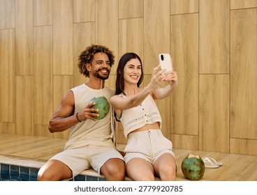 Two people enjoy coconuts while sitting in water at a pool area and using a smartphone camera  taking a selfie photo in a warm location during summer