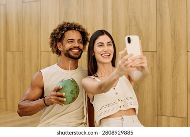 Two people enjoy coconuts while sitting in water at a pool area and using a smartphone camera  taking a selfie photo in a warm location during summer