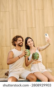 Two people enjoy coconuts while sitting in water at a pool area and using a smartphone camera  taking a selfie photo in a warm location during summer