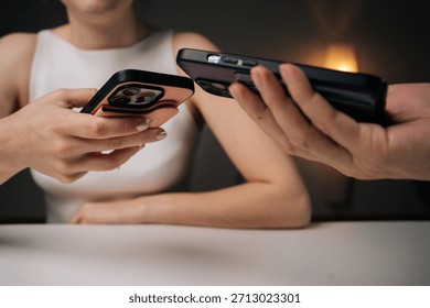 Two people exchanging data using mobile phones sitting at white table at home. Man and woman comparing smartphone data, sharing digital information in casual workspace environment.