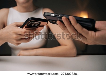 Two people exchanging data using mobile phones sitting at white table at home. Man and woman comparing smartphone data, sharing digital information in casual workspace environment.