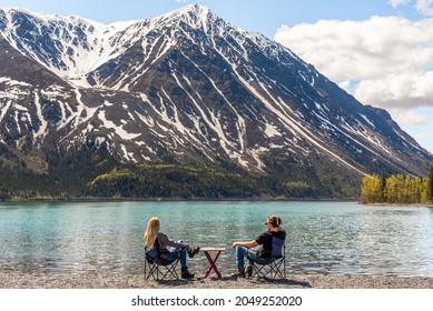 Two People, Friends, Couple Sitting On Camping Chairs With Picnic Table On Lake Shore Scenic Area In Yukon Territory, Northern Canada. Taken In Early Spring Time On Blue Sky Day. 