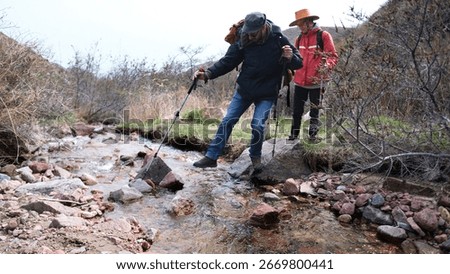 Two people go down the river. The guide is the first to cross a small river on the rocks. The tourist looks attentively at his colleague. Trekking and hiking