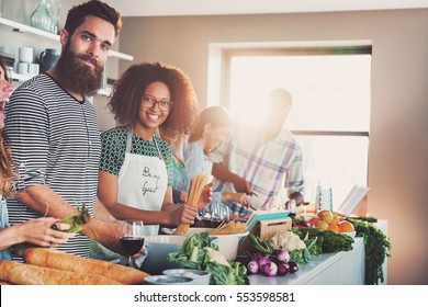 Two People From Group Of Friends Smiling At Camera While Cooking Dinner Together In Light Modern Kitchen.