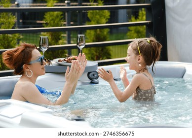 Two people are having fun in a hot tub on a sunny day. One is an adult with sunglasses, and the other is a child. They are laughing and splashing water while enjoying their time together.