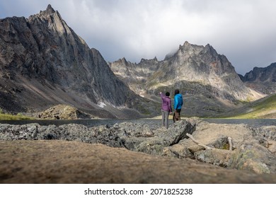 Two People, Hikers, Travellers Standing Beside Grizzly Lake In Northern Canada During Summertime Wearing Blue, Purple Clothing.
