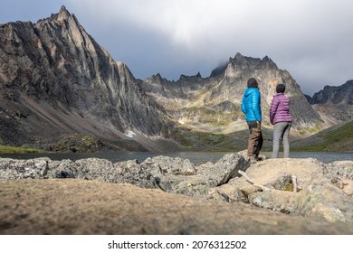 Two People, Hikers, Travellers Standing Beside Grizzly Lake In Northern Canada During Summertime Wearing Blue, Purple Clothing.
