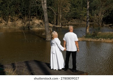 Two people hold hands while standing on a wooden dock by a lake. They are looking out at the water surrounded by green trees on a sunny day.