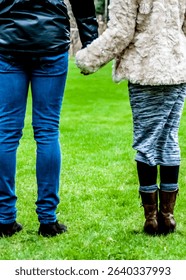 Two people are holding hands while standing on a grassy field. One person is wearing blue jeans and a black jacket, while the other is wearing a fur coat and a skirt.