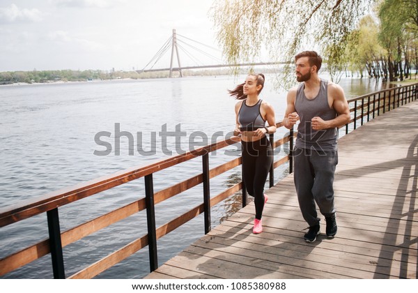 Two people\
are jogging on the bridge. There is a river nearby. Man and woman\
are looking at the river and\
running.