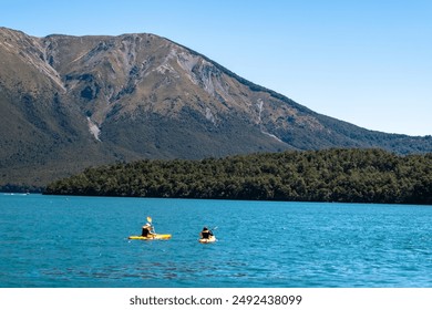 Two people kayaking on a clear blue mountain lake, surrounded by lush green forests and towering peaks under a bright blue sky. - Powered by Shutterstock