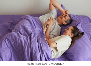 Two people are lying in bed under a purple blanket. They are laughing and enjoying their time together in a cozy and bright room during morning hours.