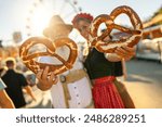 Two people at Oktoberfest or dult in Munich, holding large pretzels, smile and enjoy the festival atmosphere with a Ferris wheel in the background.