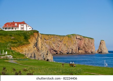 Two People On A Bench Observing The Red House On The Cliff, And Part Of The  Percé Rock(Percé (city) Quebec, Canada
