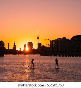 Two People On Paddle Board / Stand Up Paddler On River Spree In Berlin - Oberbaum Bridge, Tv Tower And Sunset Sky Background