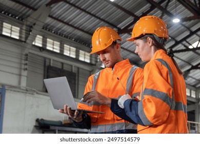 Two people in orange safety vests are looking at a laptop. They are wearing hard hats and safety gear