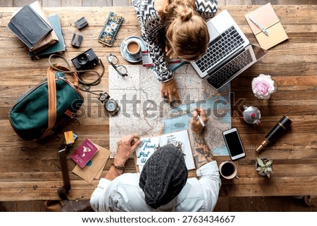 Two people planning a trip with a map and laptop on a wooden table. Includes travel essentials like a camera, coffee, and notebooks. Overhead view.