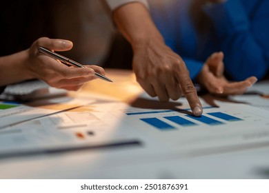 Two people are pointing at a graph on a table. One of them is holding a pen. The graph shows a downward trend, which could indicate a decrease in sales or a decline in performance - Powered by Shutterstock