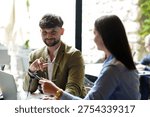 Two people are seated at a table inside a cafe. One person holds glasses and smiles while talking to the other. Their laptops are on the table as they share ideas.
