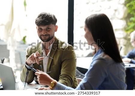 Two people are seated at a table inside a cafe. One person holds glasses and smiles while talking to the other. Their laptops are on the table as they share ideas.