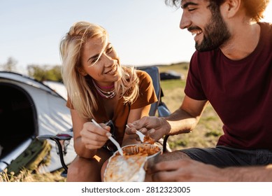 Two people share a meal using forks while sitting near a tent in a grassy area. The sun is shining and the environment is lush and green.
