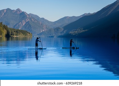 Two People In Silhouette Paddleboarding Across Calm Lake