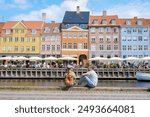 Two people sit by the canal in Copenhagen, enjoying the sunshine and the view of colorful buildings. Copenhagen Denmark 