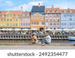 Two people sit on a cobblestone walkway along a canal in Copenhagen, enjoying the sunshine and a view of colorful buildings. Copenhagen Denmark