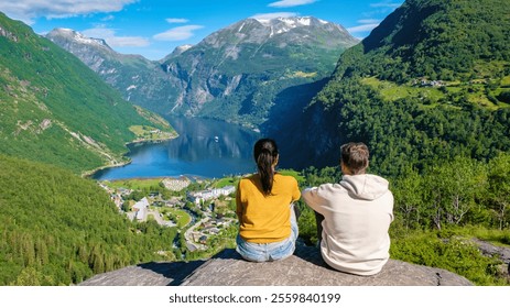 Two people sit on a rocky ledge overlooking a stunning Norwegian fjord, surrounded by lush green mountains and a clear blue sky. Geiranger Fjord Norway. A diverse couple of men and women on vacation - Powered by Shutterstock